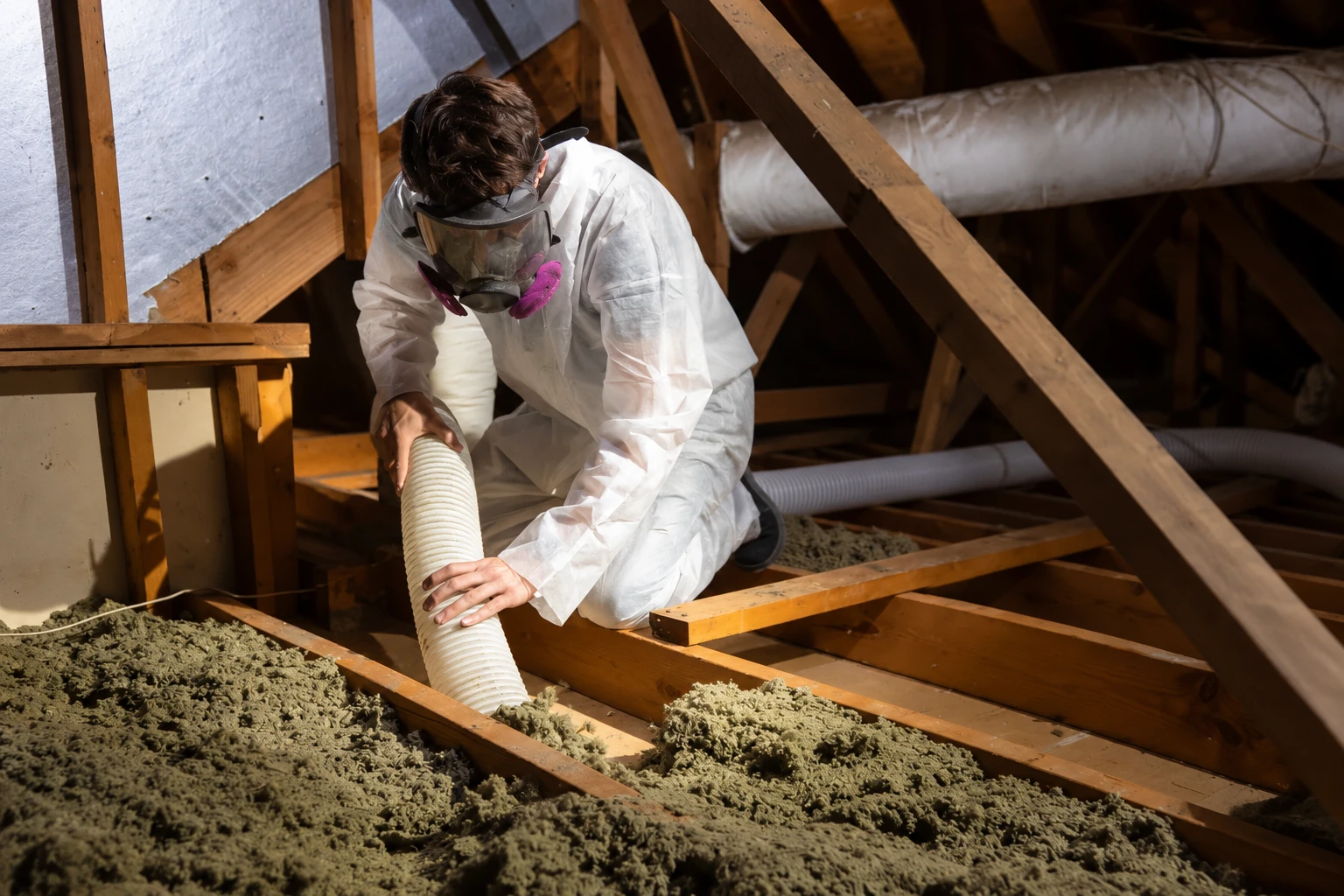 A worker in protective coveralls and a respirator uses a hose to remove loose insulation from an attic floor.